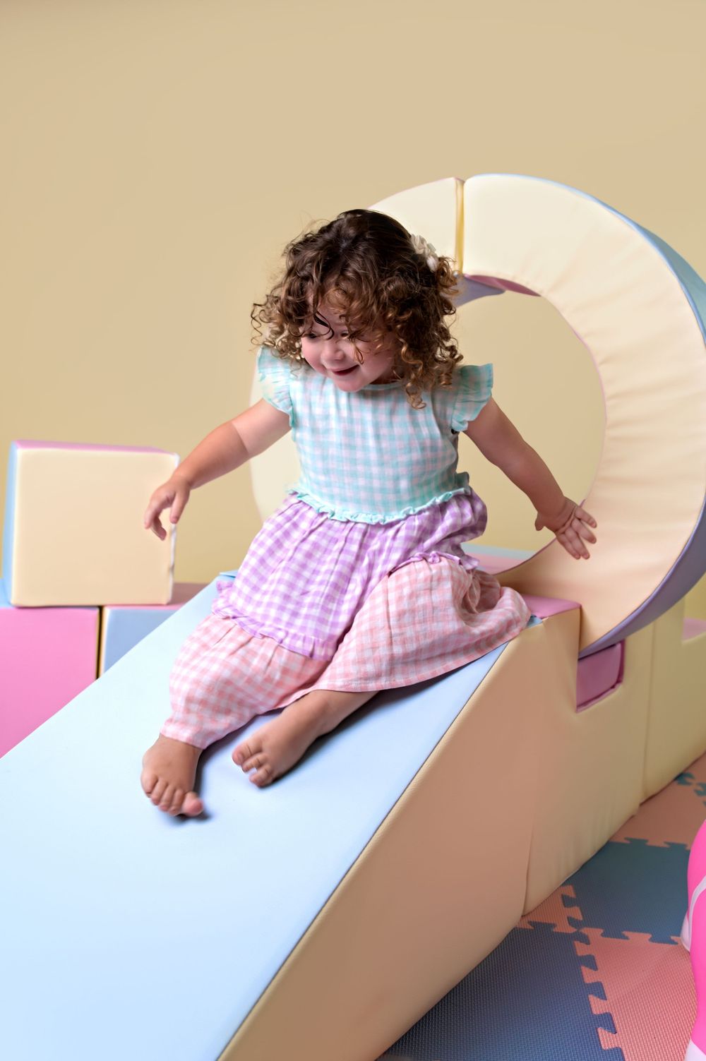 Small child sliding down blue slide wearing pink gingham dress and light blue top with white frills.