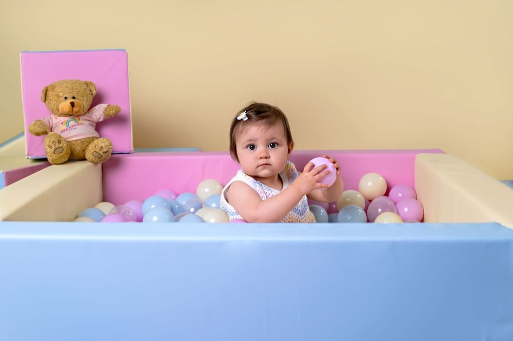 Baby playing in a blue and pink playpen filled with toys and a teddy bear.