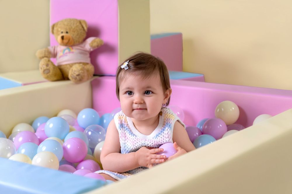 Baby in ball pit with colorful pastel balls and teddy bear against beige background.