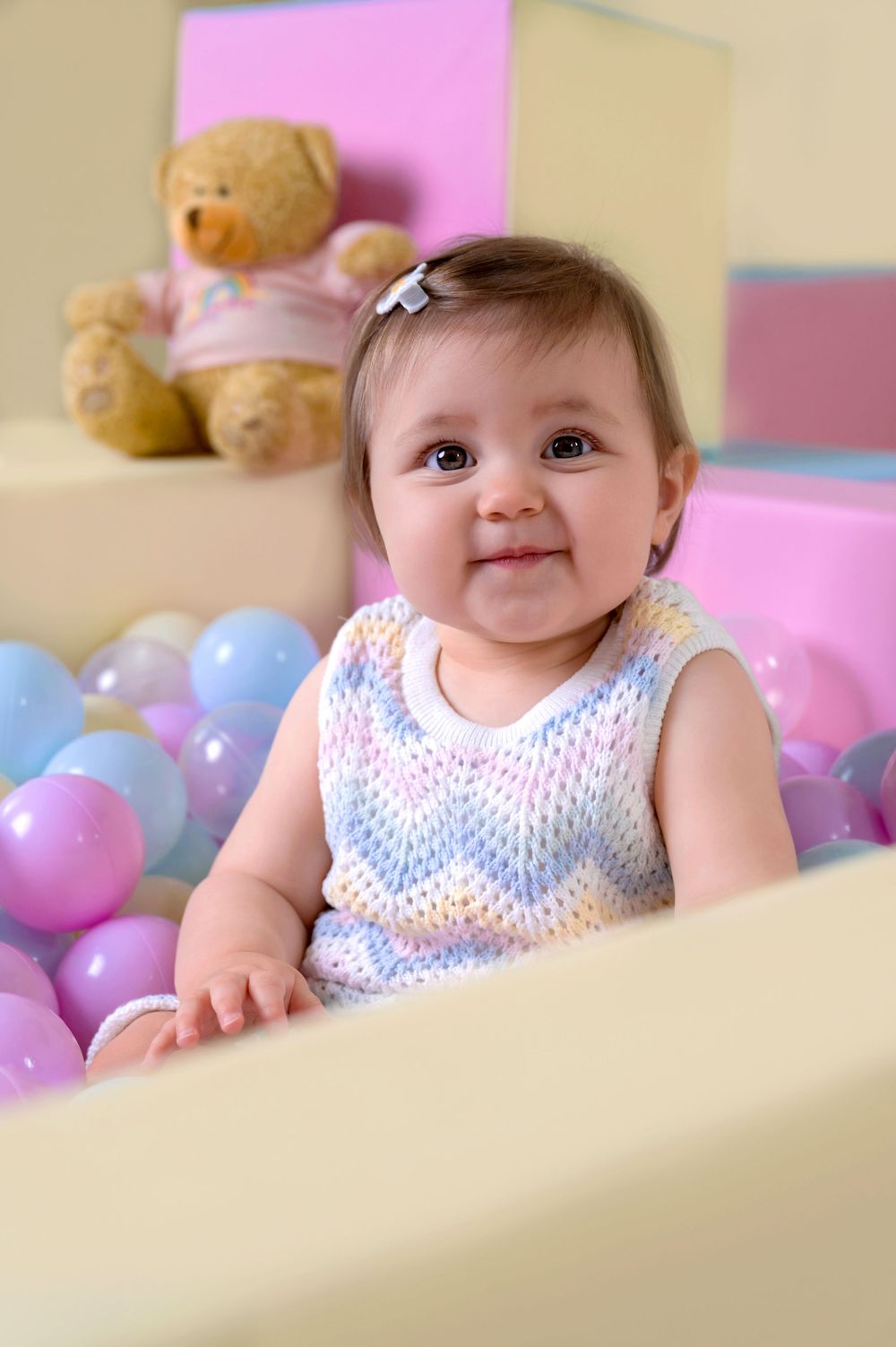 Baby in chevron pattern dress plays among pink and blue balloons with teddy bear in background.