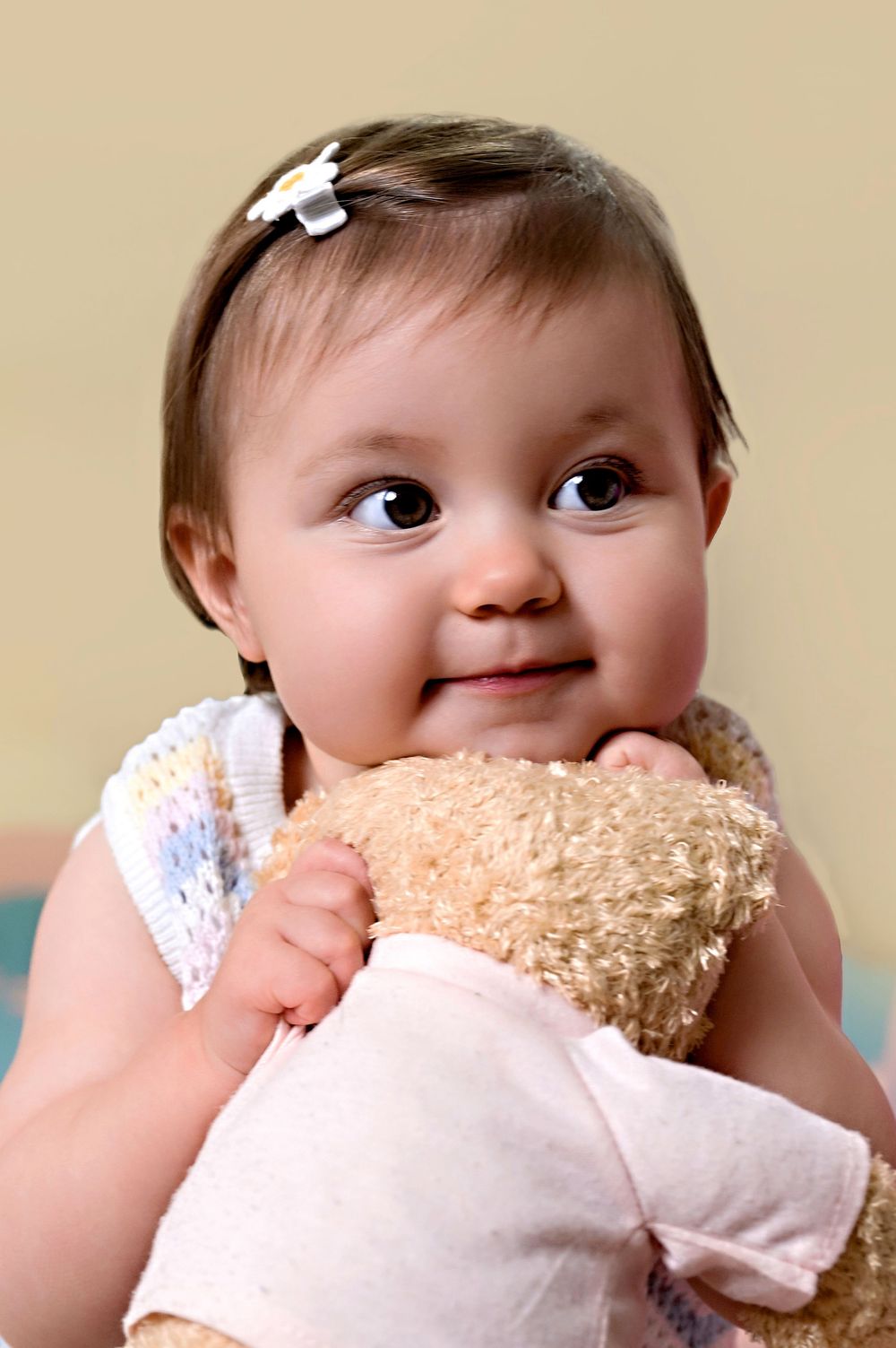 Baby in white dress hugging soft teddy bear against neutral background.