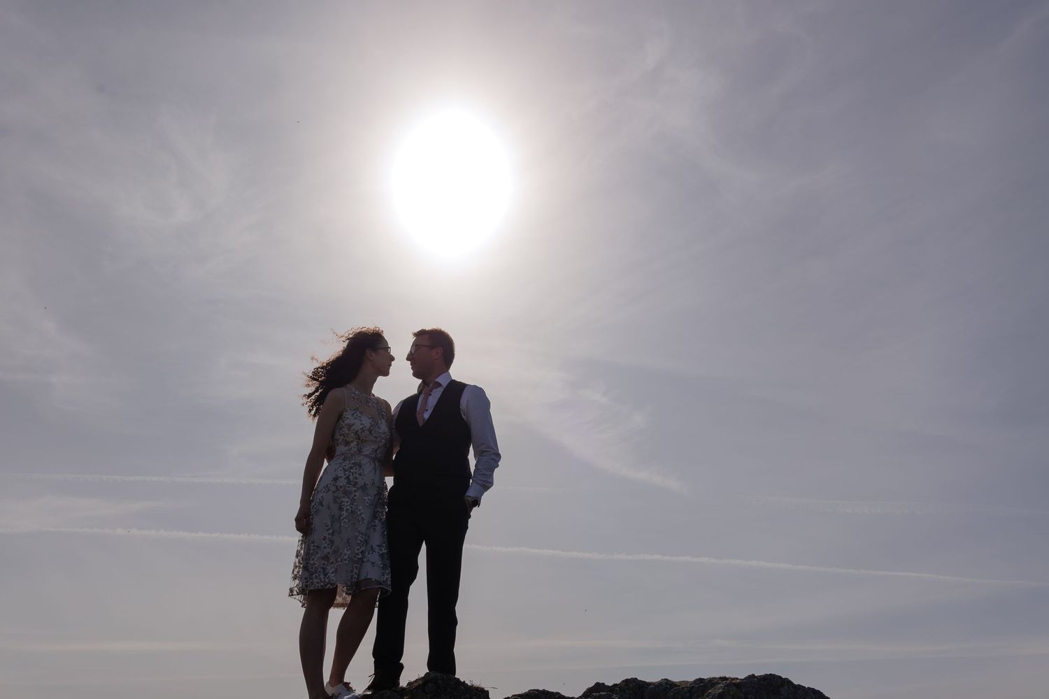 A silhouetted couple stands together on rocky terrain against a bright sun and cloudy sky backdrop.