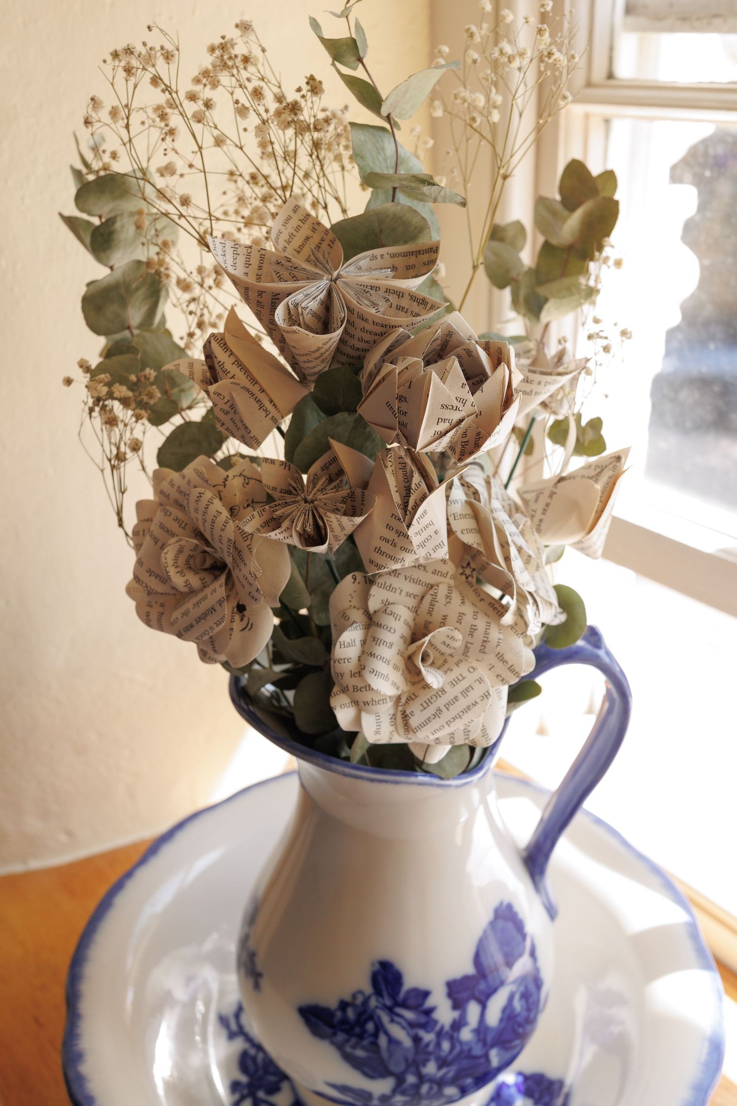Paper flowers and dried baby's breath arranged in a blue and white porcelain pitcher vase on a windowsill.