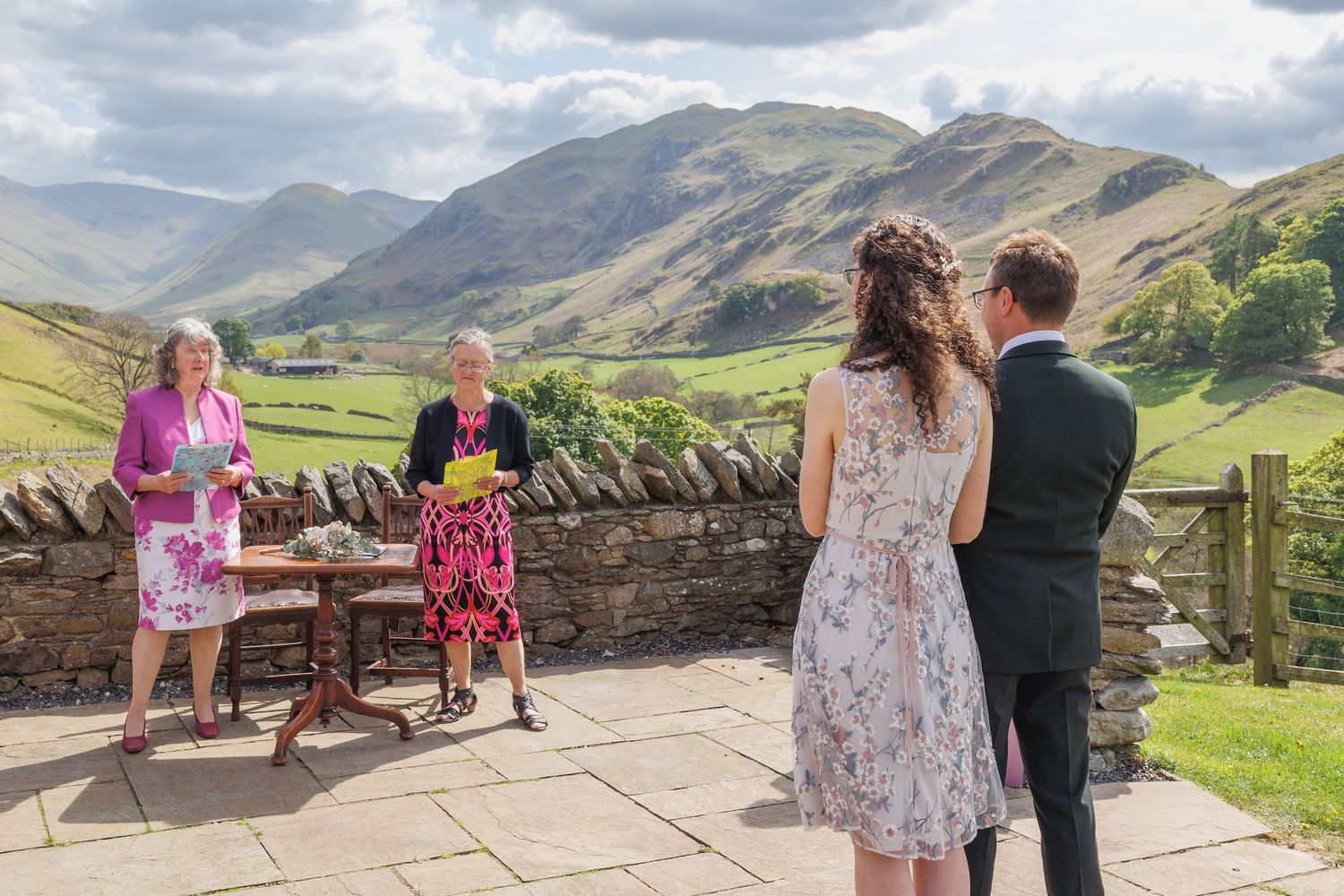 Two people enjoying the scenic mountain views from a stone wall terrace overlooking a vast green valley in the Lake District.