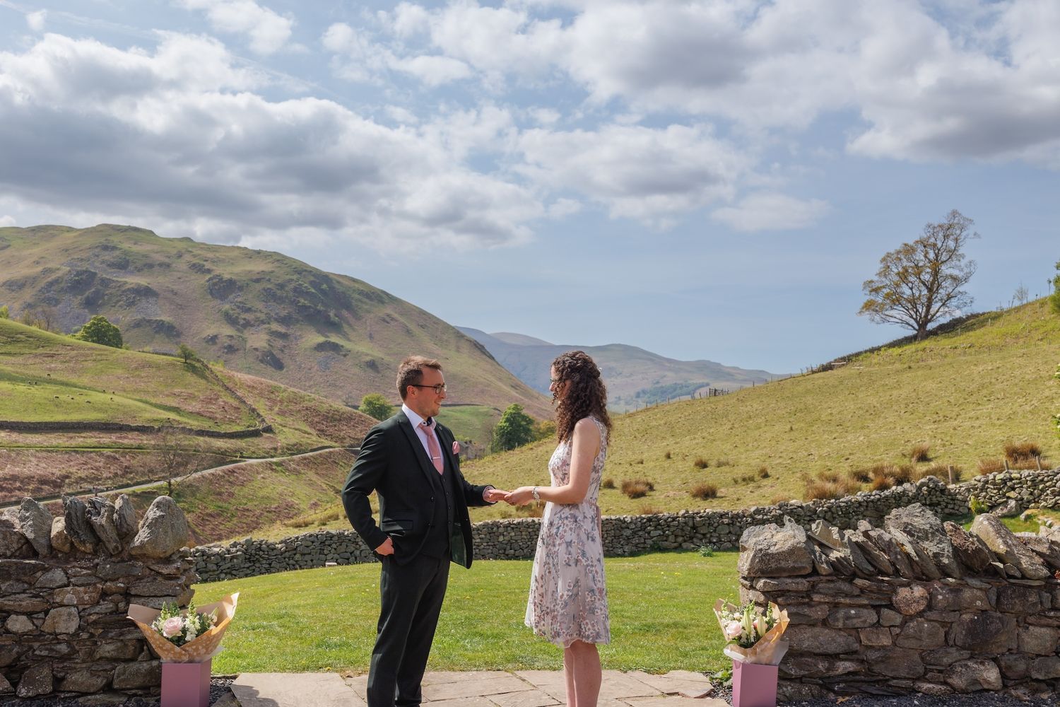 A couple shares a moment together in front of a scenic mountainous landscape with stone walls and green hills.