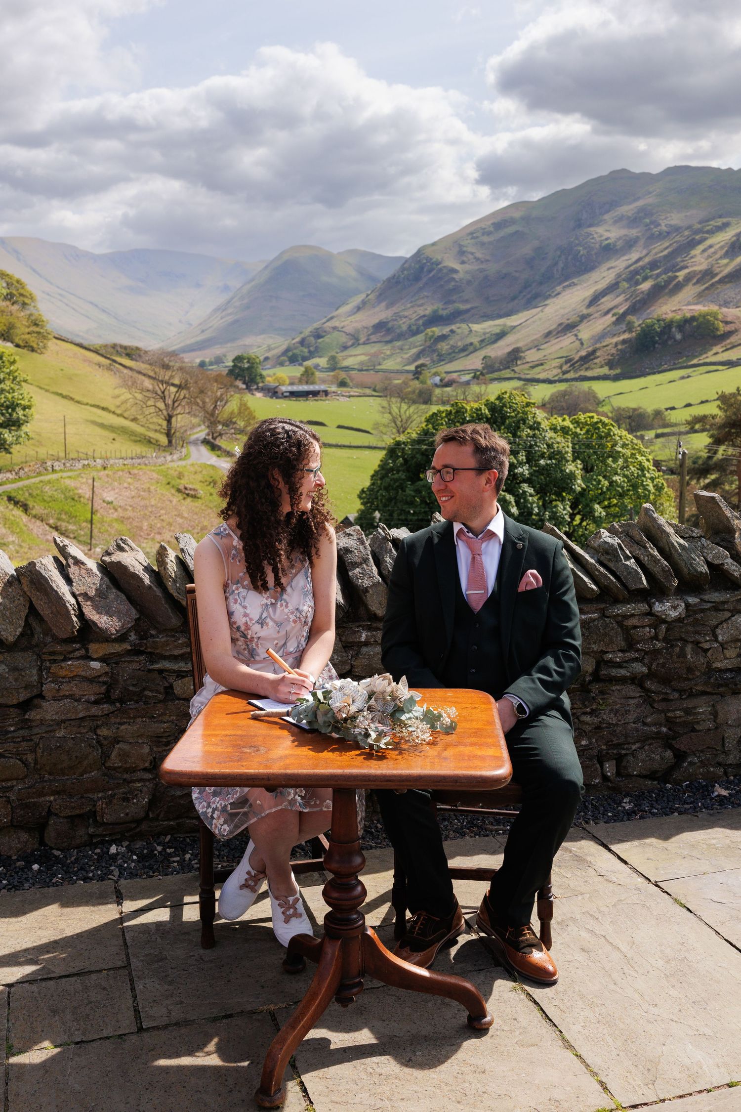 A couple sits together on a stone wall bench overlooking scenic green mountains and valleys in the Lake District.