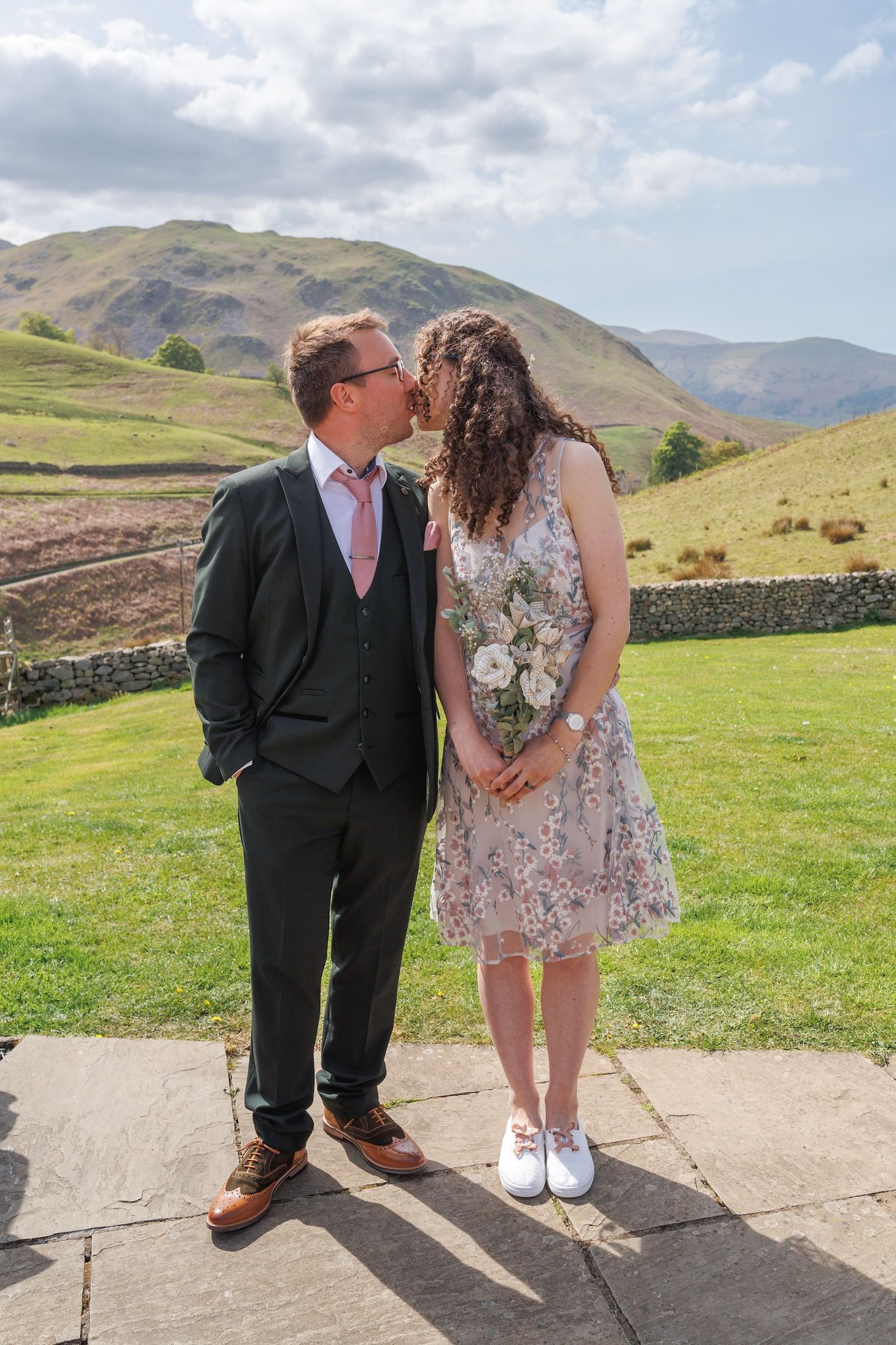 A couple in wedding attire poses playfully on a stone path with rolling green hills and mountains in the background.