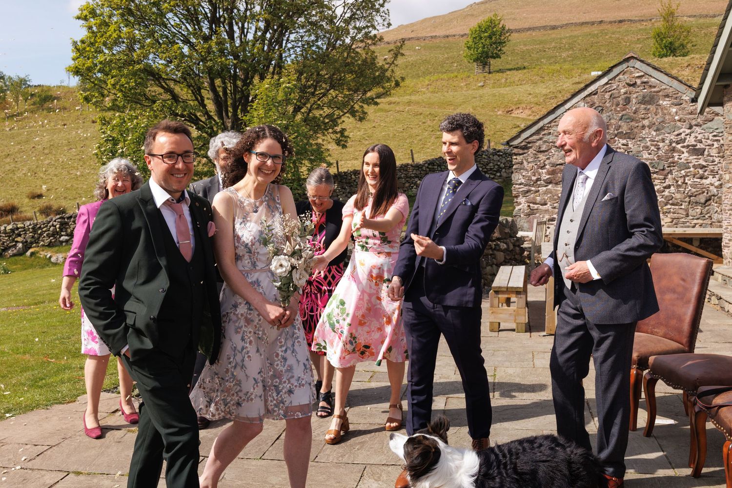 A small group gathers outdoors on a sunny day near a stone wall, with rolling hills visible in the background.