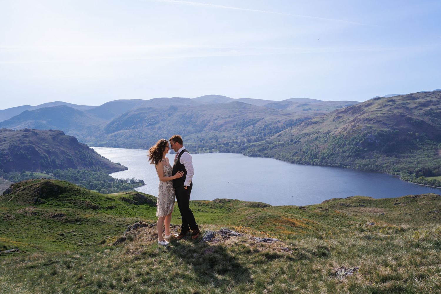 A couple enjoys a romantic moment overlooking a scenic mountain lake with rolling hills and blue water in the backdrop.