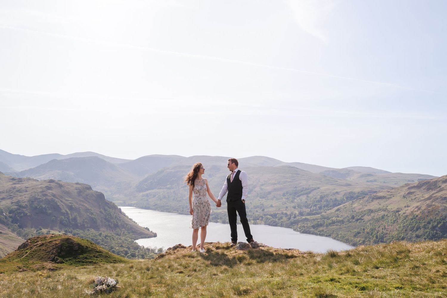 Two people stand together on a grassy hilltop overlooking a scenic lake and mountain landscape in Scotland.