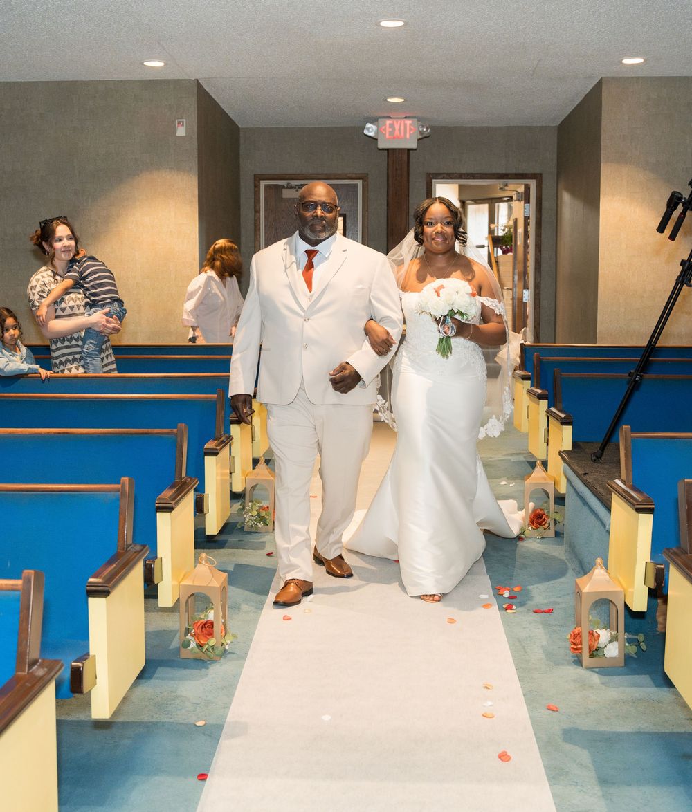 A wedding ceremony in a chapel with cream-colored pews and white aisle runner decorated with flower petals.