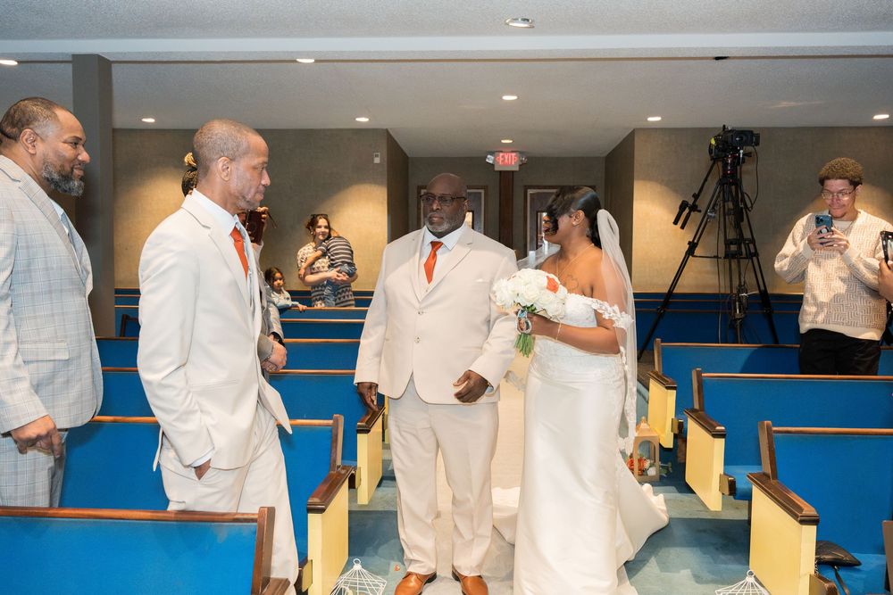 A wedding ceremony group performs a line dance on a blue floor while wearing all white attire.