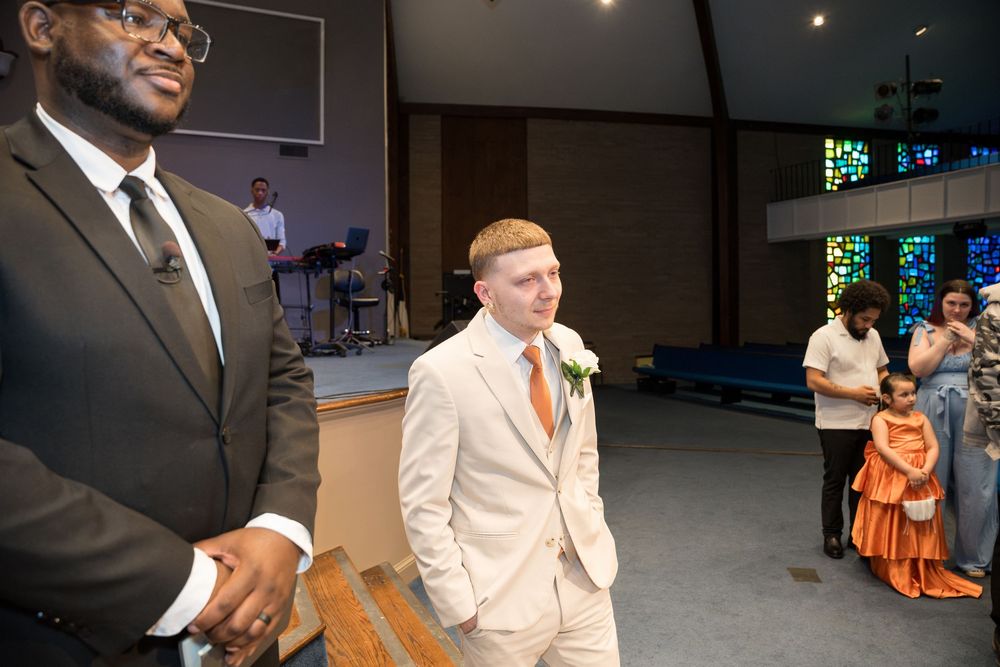 A groom in a light colored suit stands near someone in a dark suit at what appears to be a church ceremony.