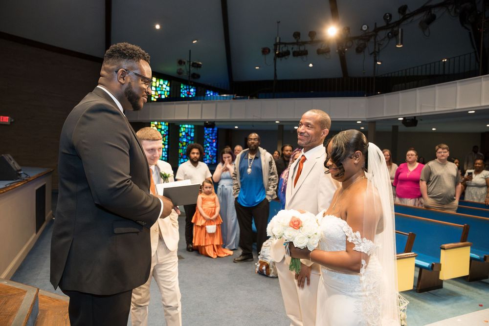 An indoor wedding ceremony takes place in a modern church or chapel space with soft lighting and gathered guests.