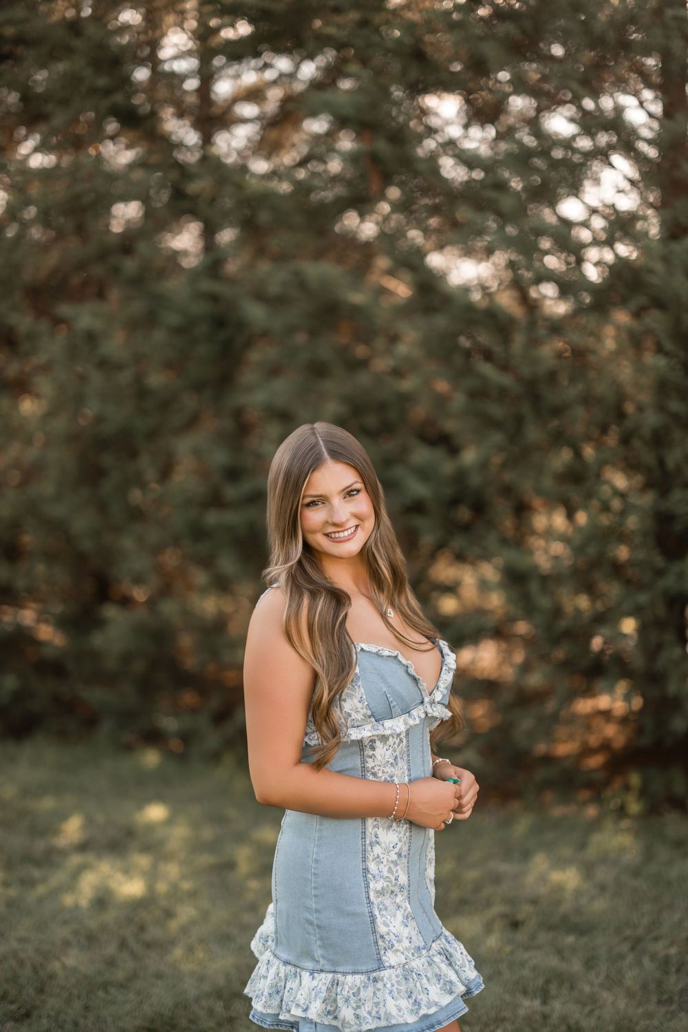 A person wearing a denim and floral ruffle top poses outdoors against a natural green background during golden hour.