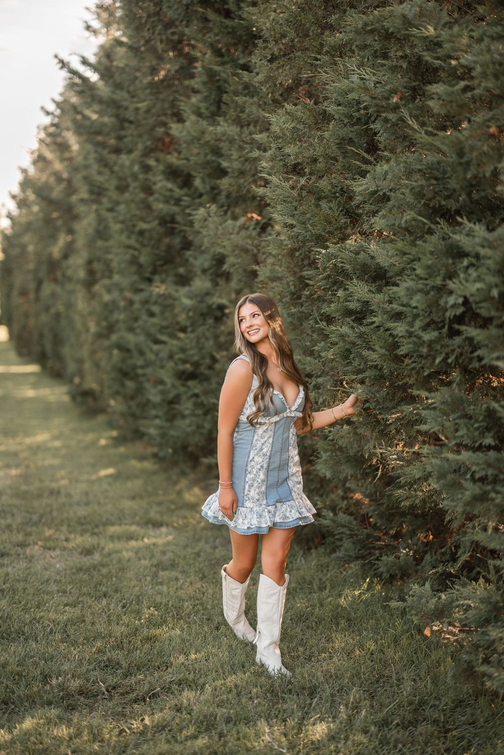 A woman in a floral dress and white cowboy boots poses near evergreen trees in golden evening light.