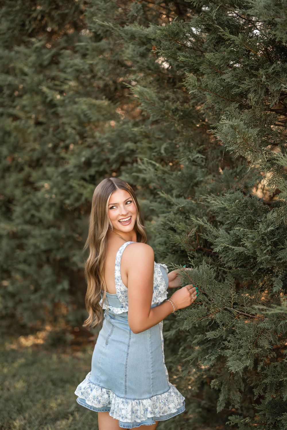A young woman in a denim and floral ruffle dress stands beside evergreen bushes in a garden setting.