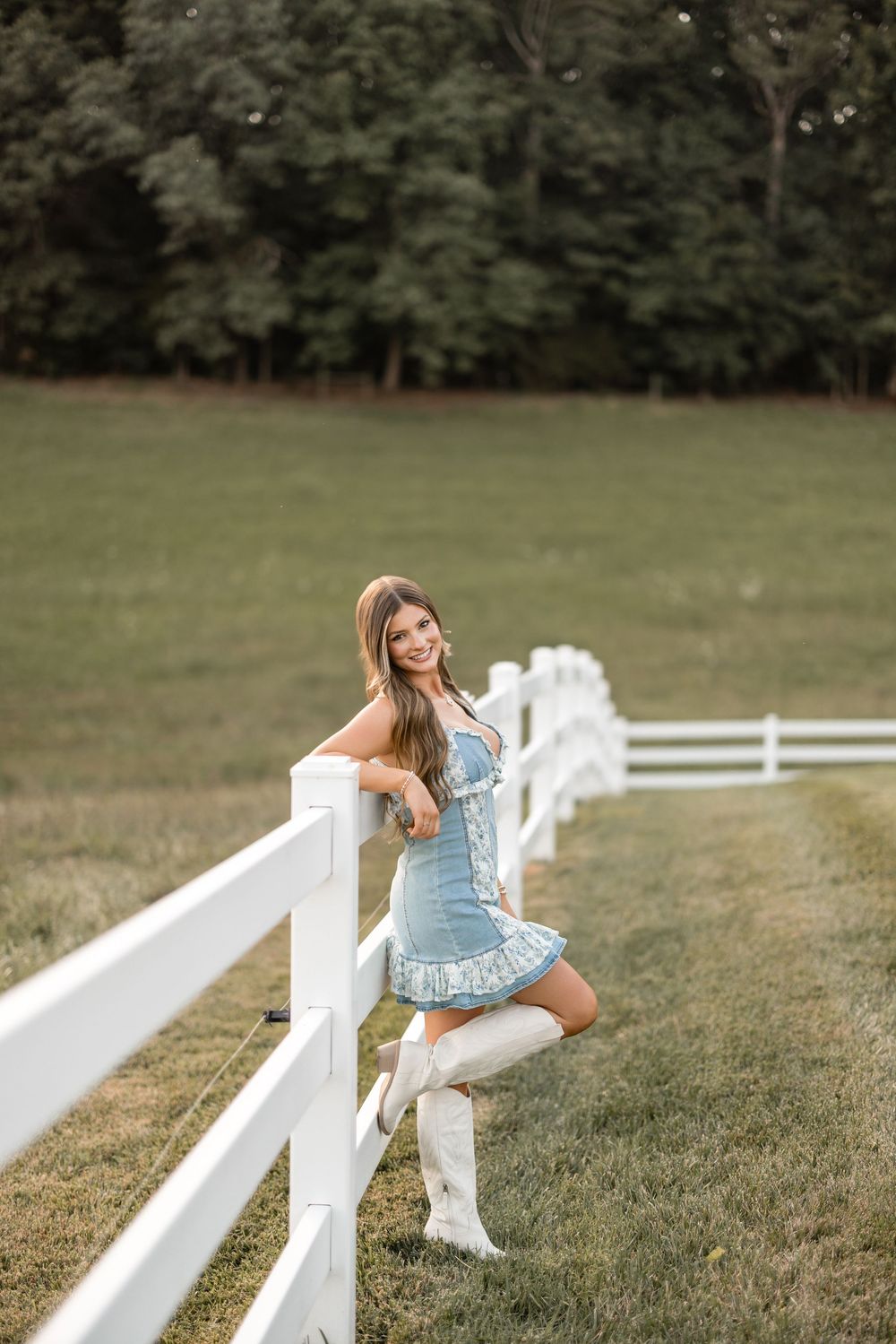 Person in light blue dress and white boots poses playfully along a white fence on grassy field with trees in background.