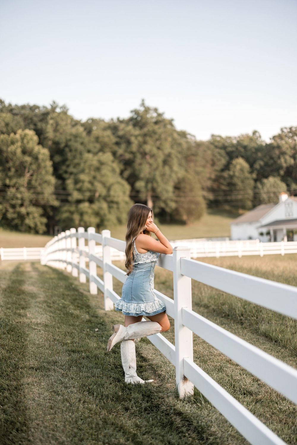 A small child in blue sits on a white wooden fence with trees in the background.