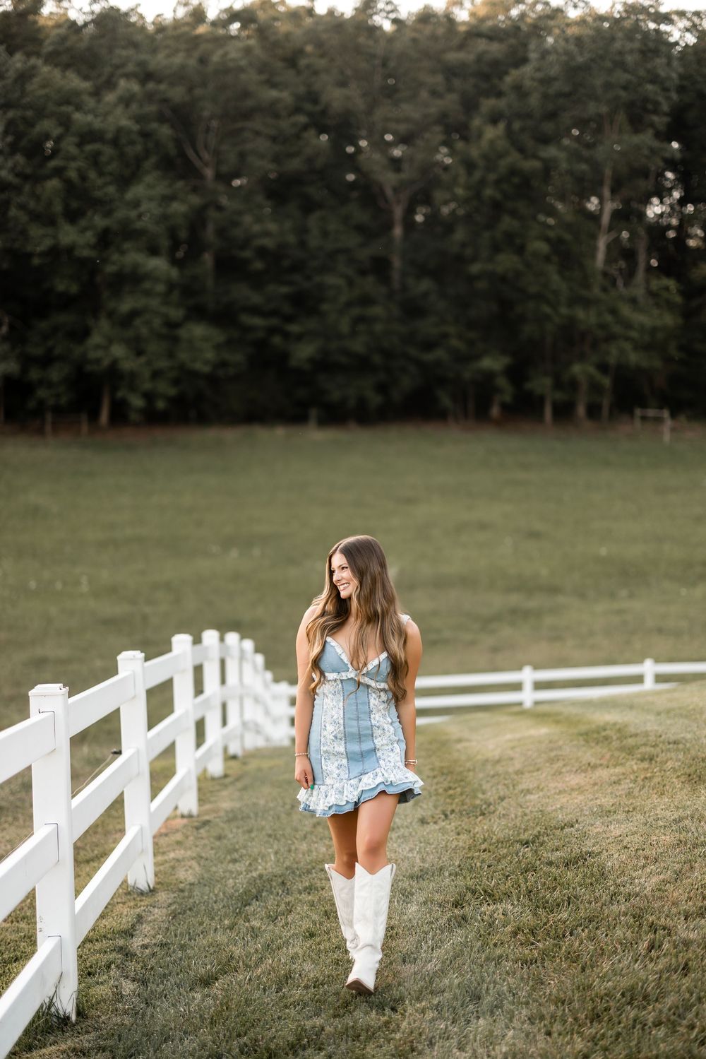 A serene country scene of a white wooden fence running along a grassy field at sunset with scattered trees in background.