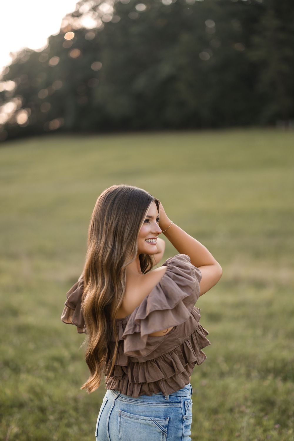 A person wearing a ruffled brown top and jeans stands in a sunlit field during golden hour.