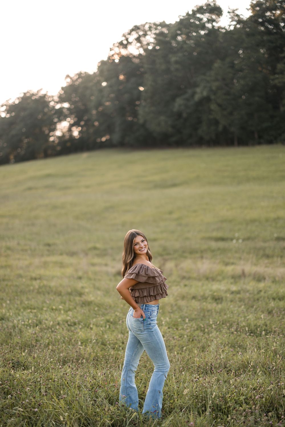 Person in casual attire poses against sunset in grassy field wearing brown top and light denim jeans.