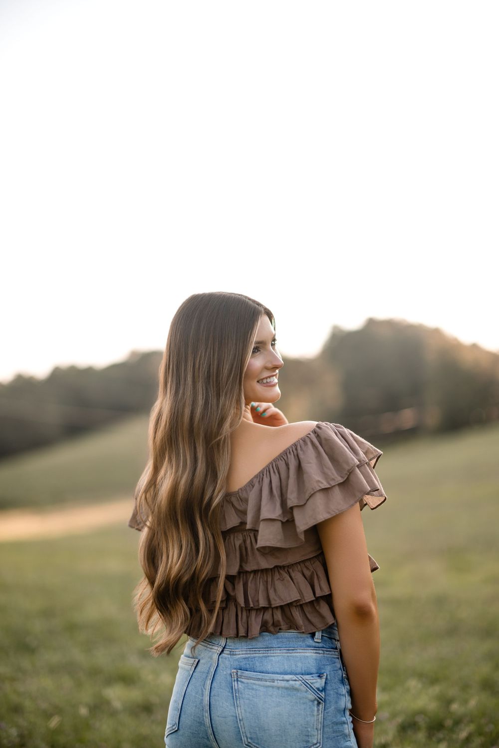 Someone with long wavy hair wearing a brown ruffled top and jeans stands in a sunny field at sunset.