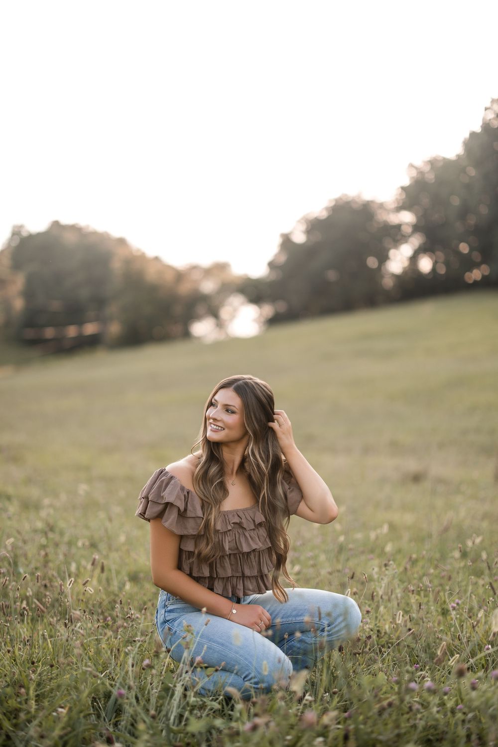 Person wearing a ruffled top and jeans lounges in a sunny meadow with a joyful expression at sunset.