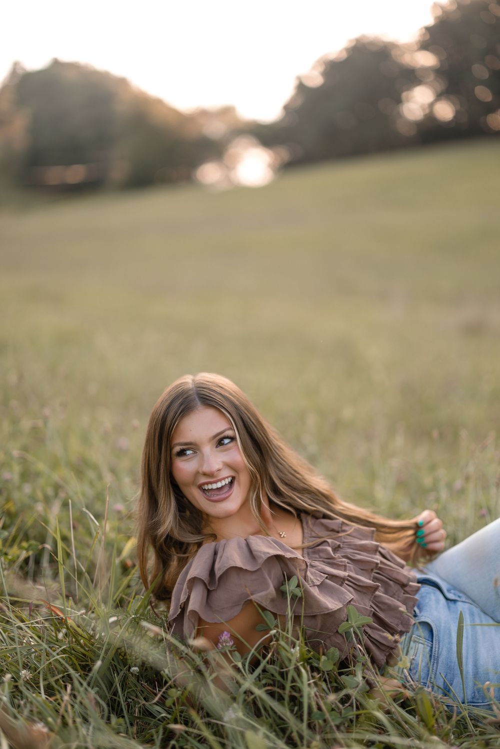 Someone relaxes in a grassy field wearing a ruffled brown top and jeans during golden hour.