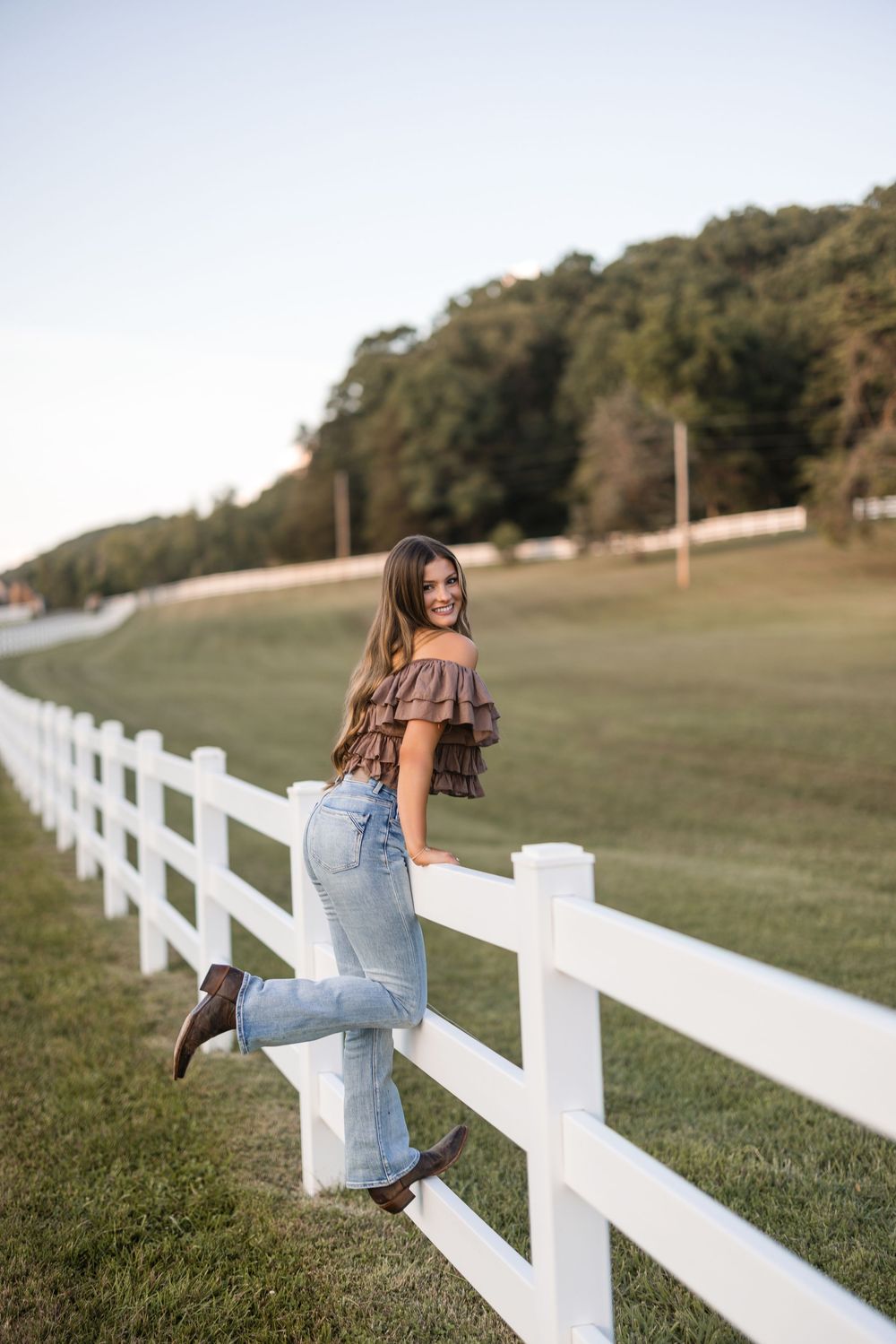 Woman in ruffled top and jeans poses casually by a white fence on a sunny day at a rural farm property.