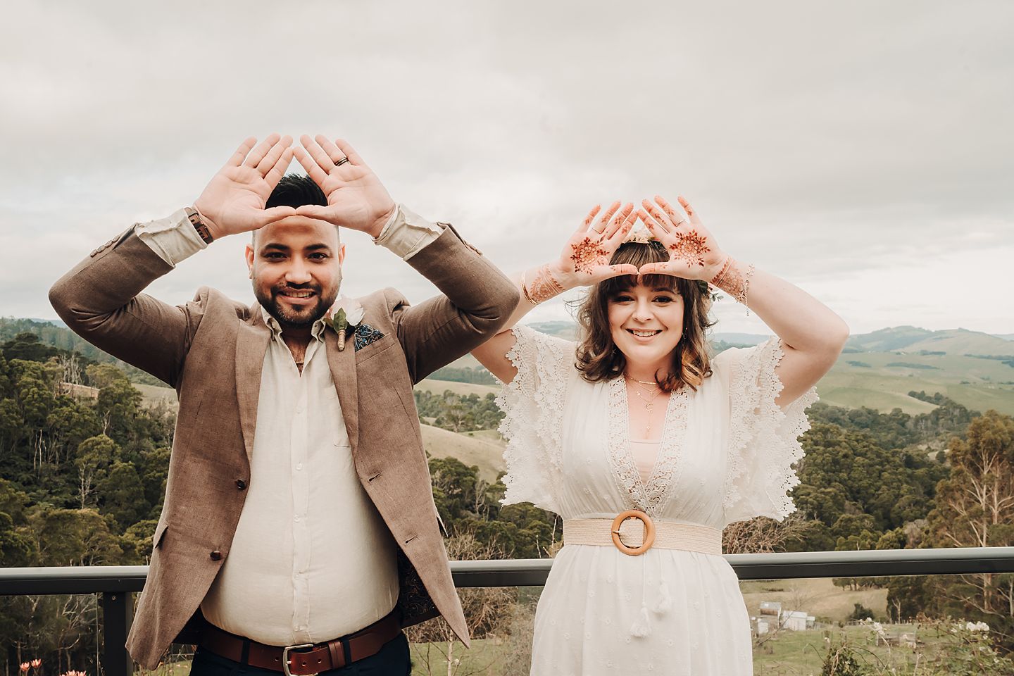 A smiling couple makes heart shapes with their hands against a scenic mountain landscape background.