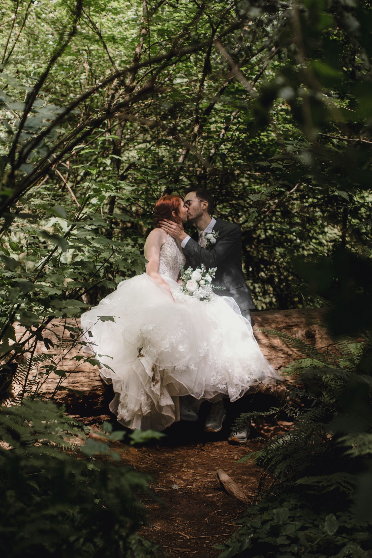 A romantic wedding photo of a couple embracing on a woodland path surrounded by lush green foliage.