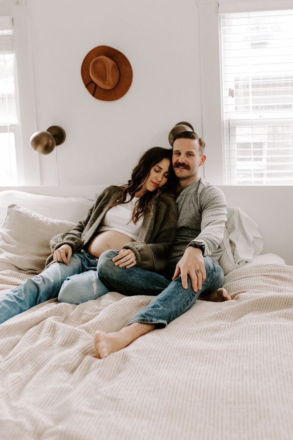 An intimate maternity portrait of a couple relaxing on white bedding with a brown hat on the wall.