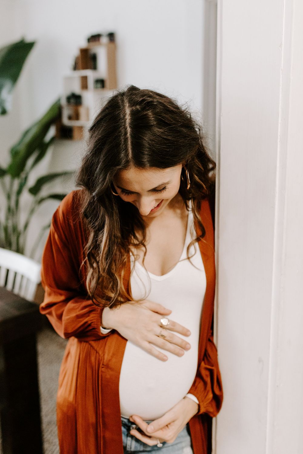 A serene maternity portrait of a woman in rust-colored cardigan touching her pregnant belly.