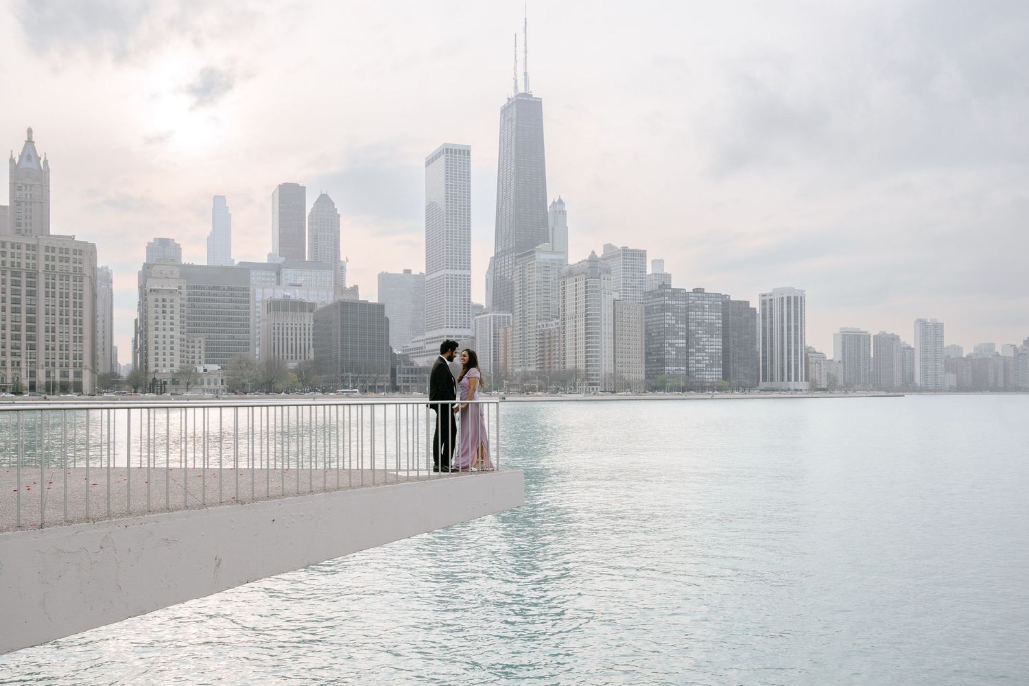 A couple stands at a railing overlooking Lake Michigan with Chicago's iconic skyline in the background on a misty day.