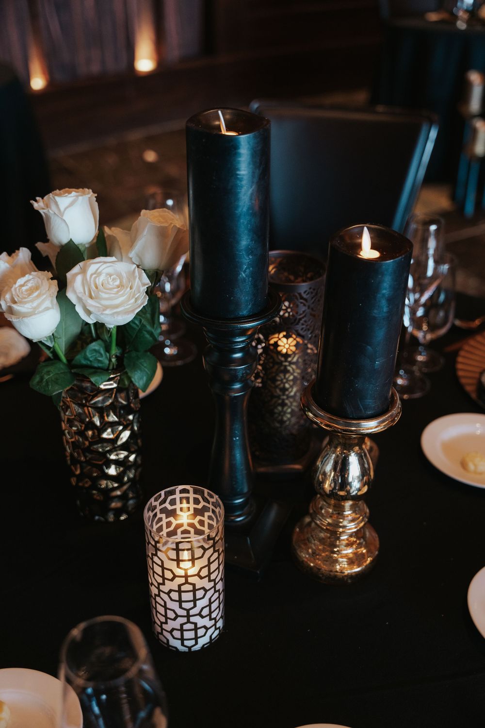 Elegant table setting with black candles, white roses and decorative metallic votives creating a moody romantic atmosphere.
