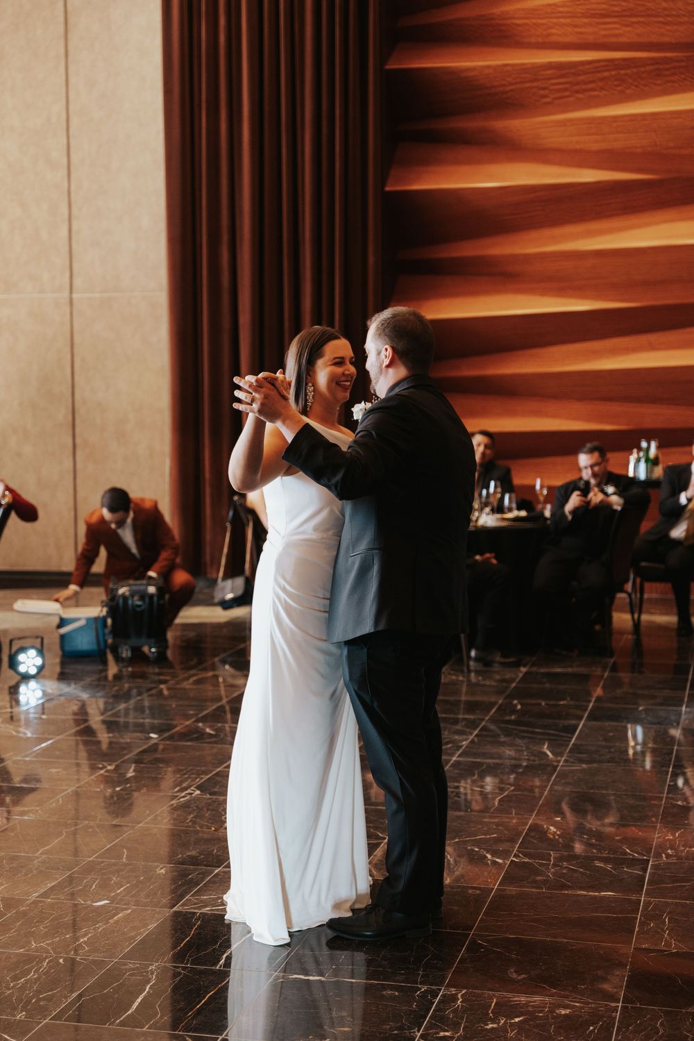 A couple dances together on a polished marble floor with warm wood paneled walls in the background.