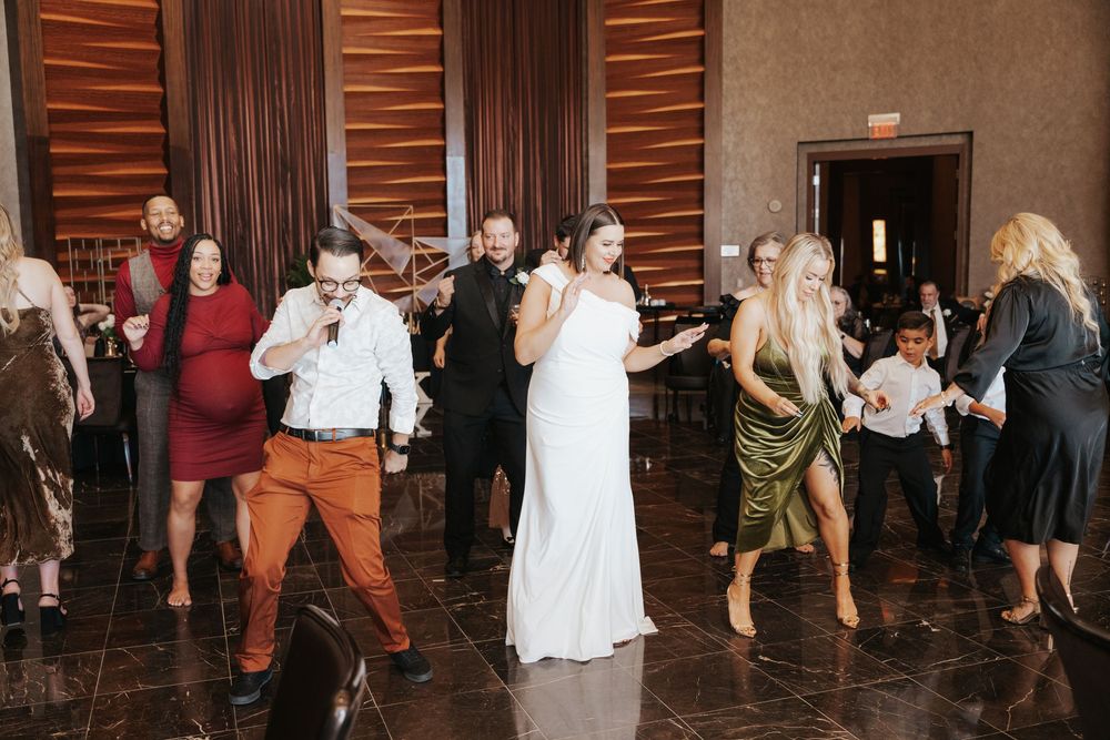 Wedding party guests dance together on a polished wooden dance floor at an indoor reception venue.