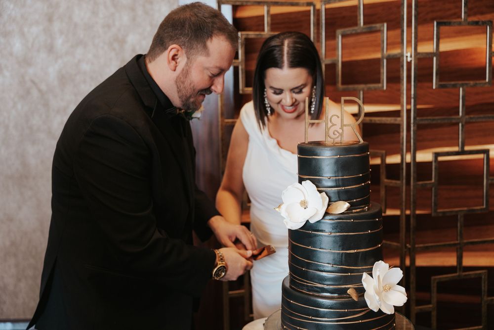 A couple cuts into an elegant black tiered wedding cake decorated with white floral accents at their reception.