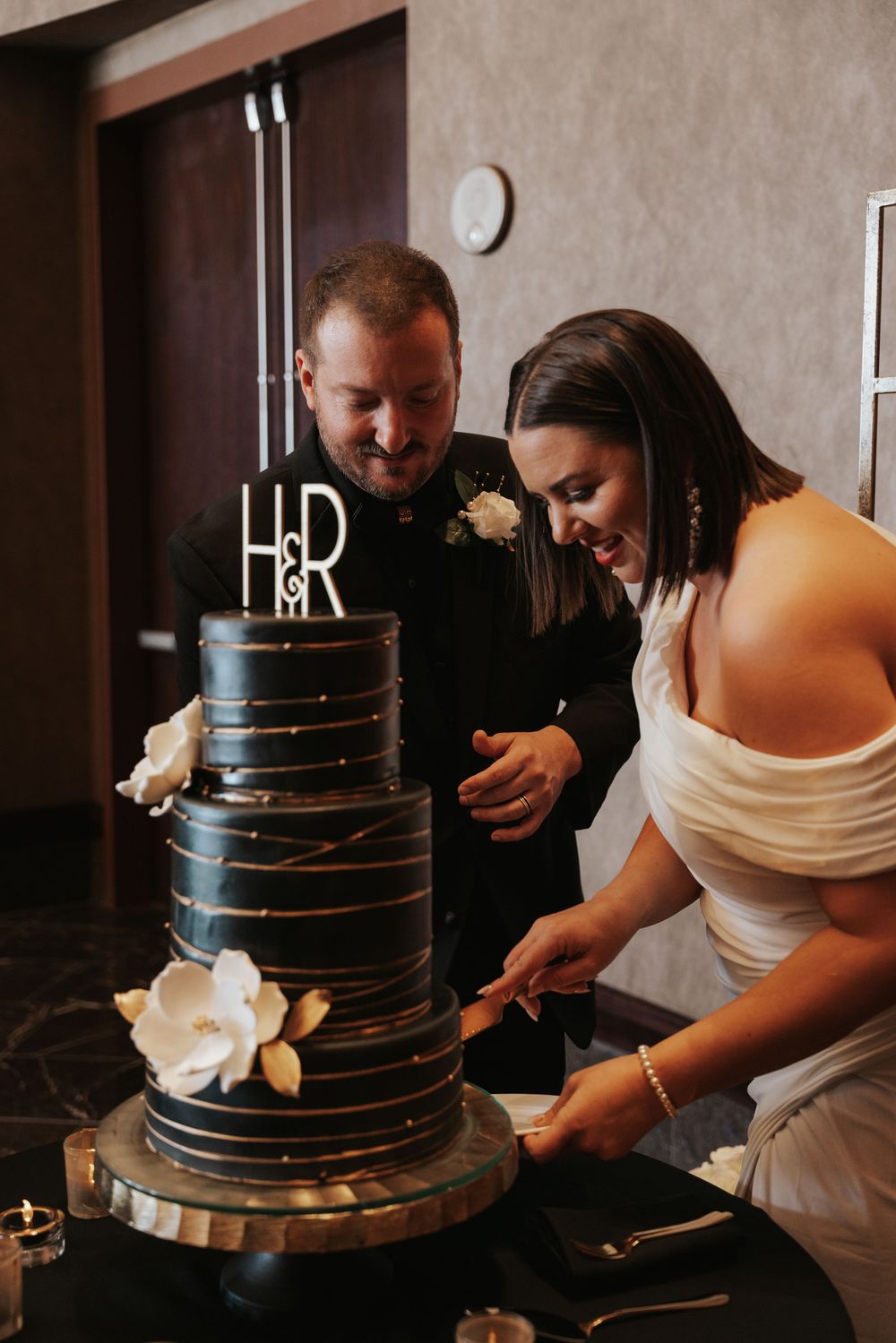 Newlyweds cut into their elegant black tiered wedding cake decorated with marble details and topped with H&R initials.