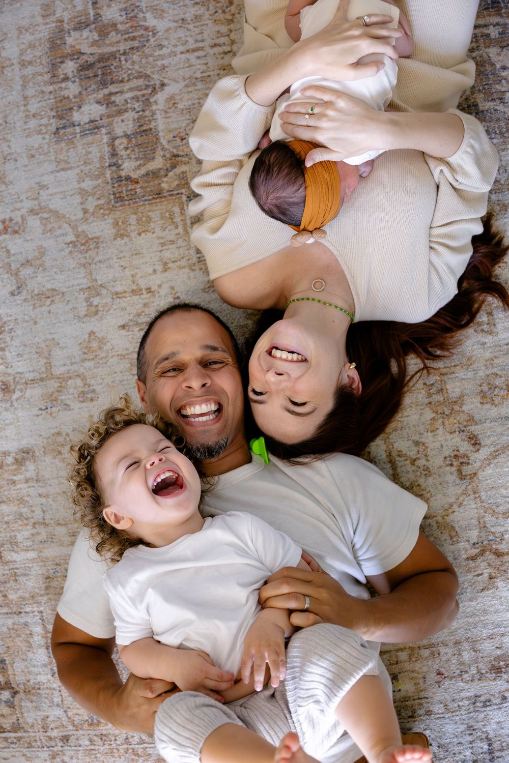 A joyful family of three laying together on a rug, laughing and embracing while wearing casual light-colored clothing.