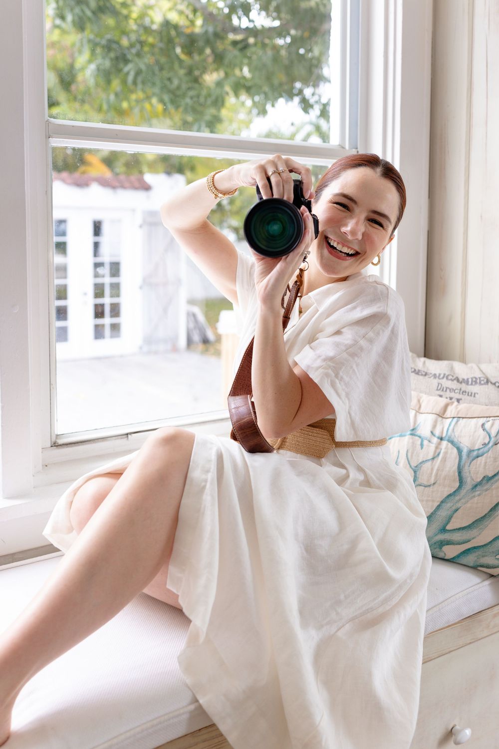 A photographer in a white dress sitting by a window holding a camera and smiling brightly.