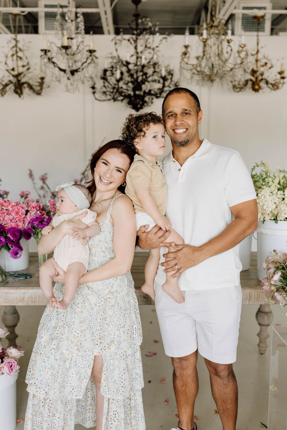 Family wearing coordinated white outfits poses together against elegant backdrop with crystal chandeliers.