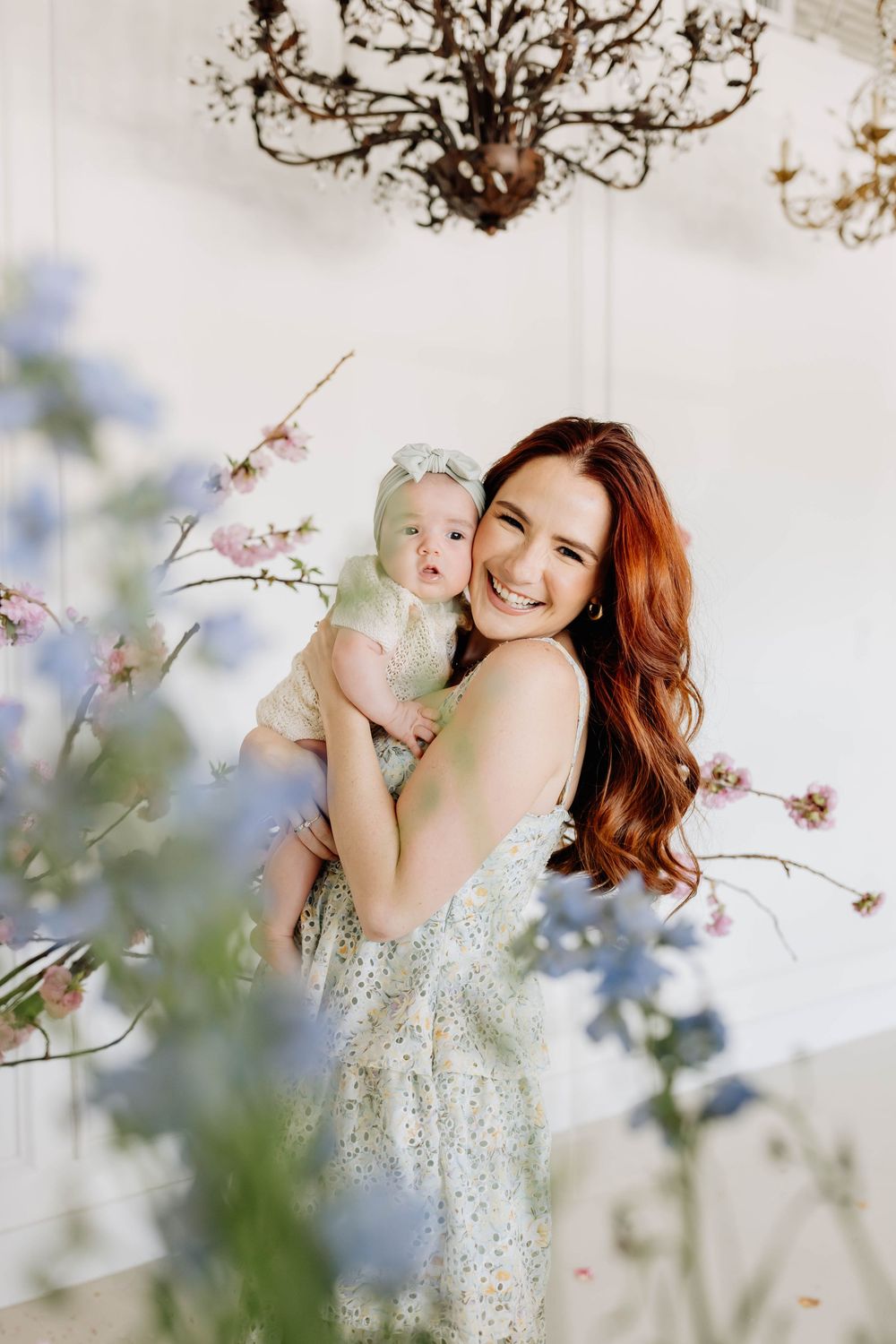A woman in a sparkly dress holds a baby against a dreamy floral backdrop with cherry blossoms and soft lighting.