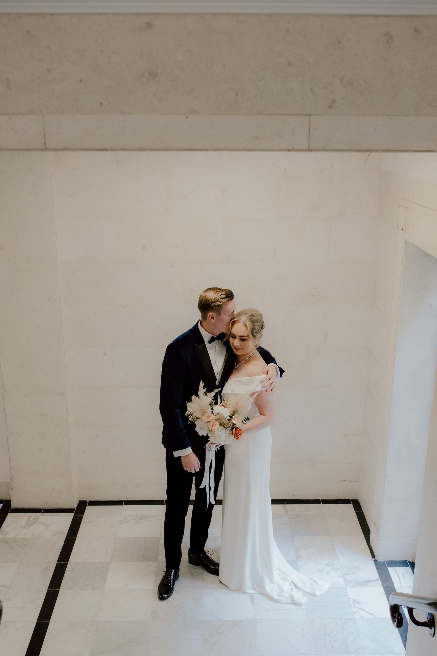Elegant couple in classic black tuxedo and white wedding gown pose together holding a delicate bouquet against white walls.