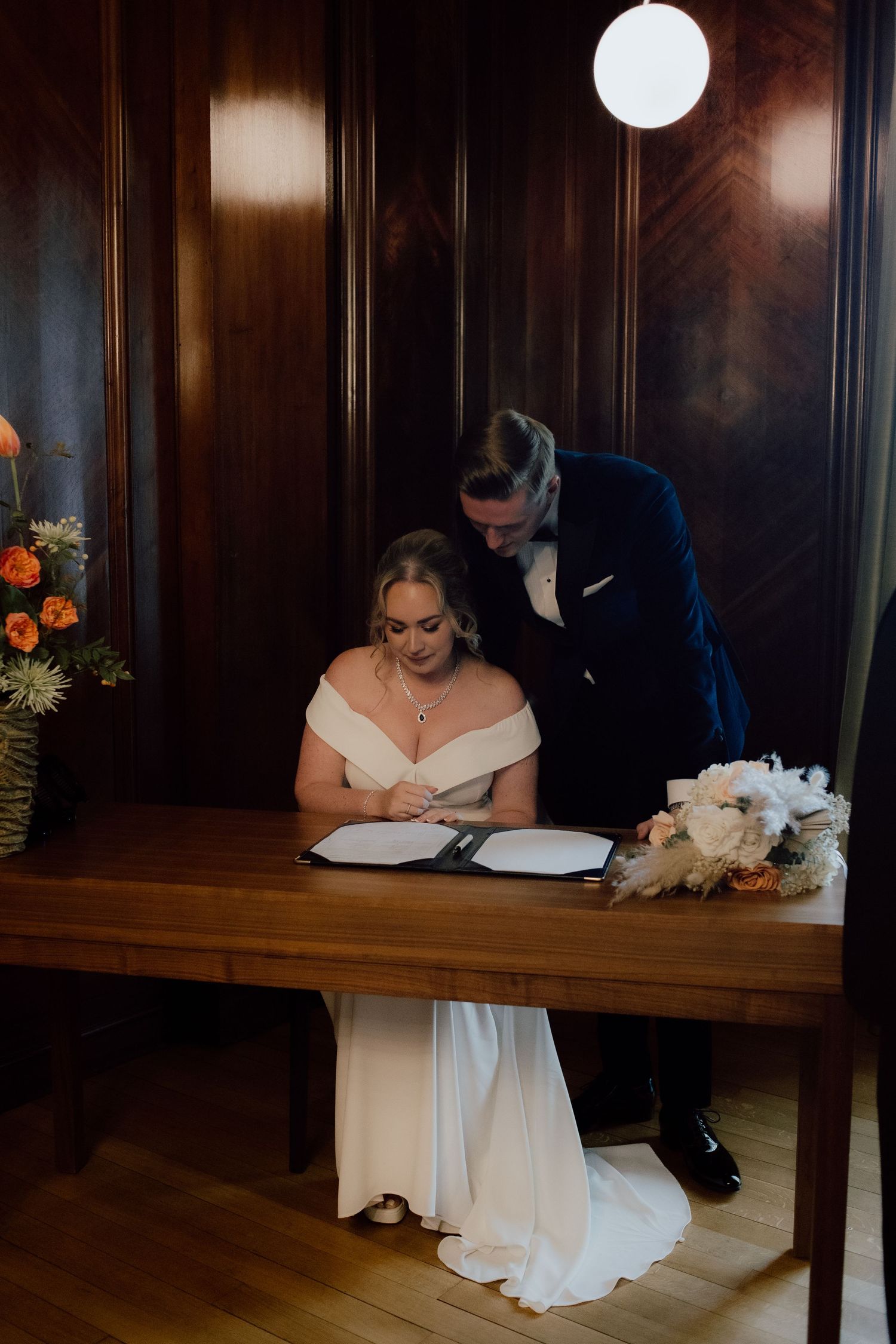 A couple signing their marriage documents at a wooden table with a white floral bouquet in an elegant wood-paneled room.