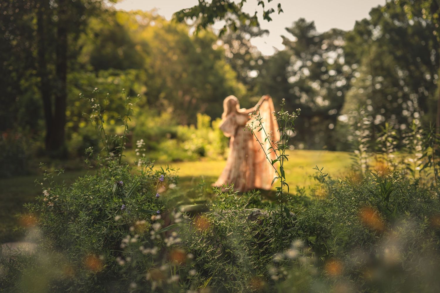 Someone in a flowing pink dress stands in a dreamy garden setting backlit by golden sunset light through the trees.