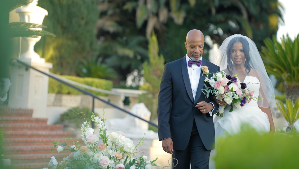 A newlywed couple stands together on outdoor stone steps surrounded by floral arrangements on their wedding day.