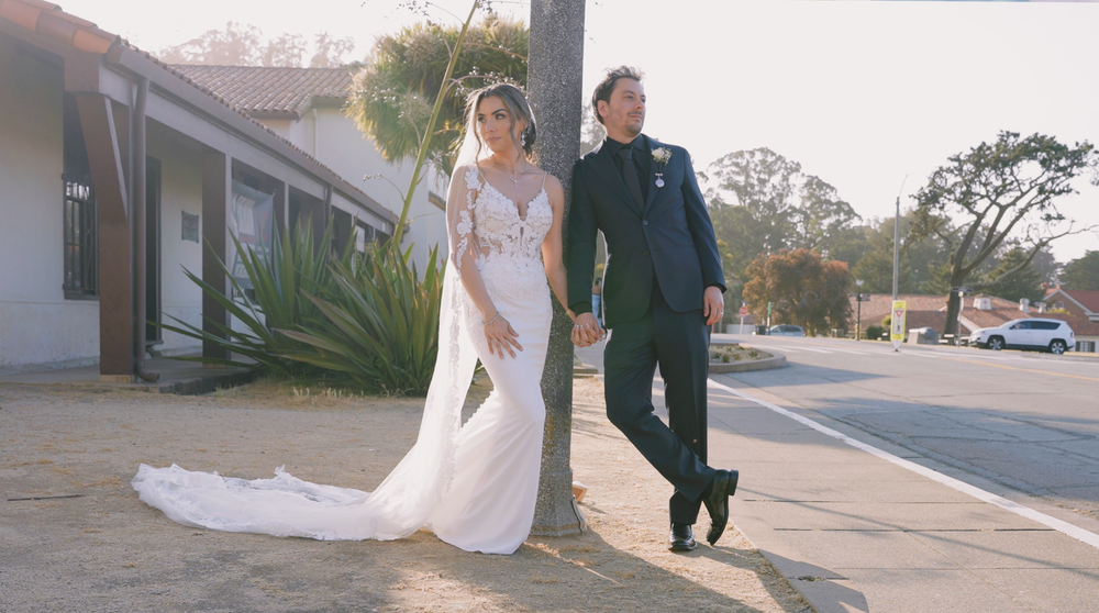A couple in wedding attire poses casually against a building exterior.