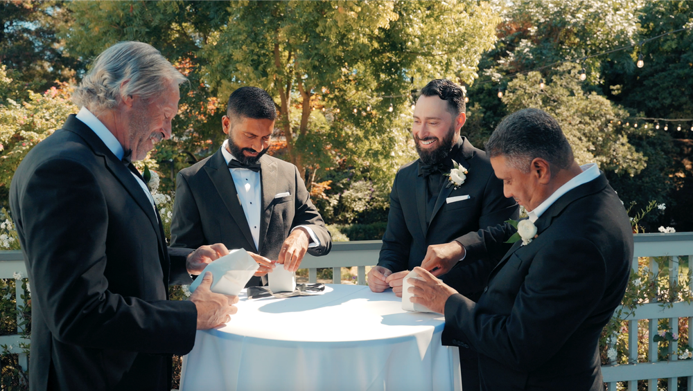 A group of formally dressed people gather around a cocktail table at an outdoor wedding reception.